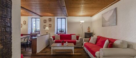 Una sala de estar acogedora con techo de madera, un sofá blanco con cojines rojos, una mesa de centro blanca y un comedor con una silla roja.