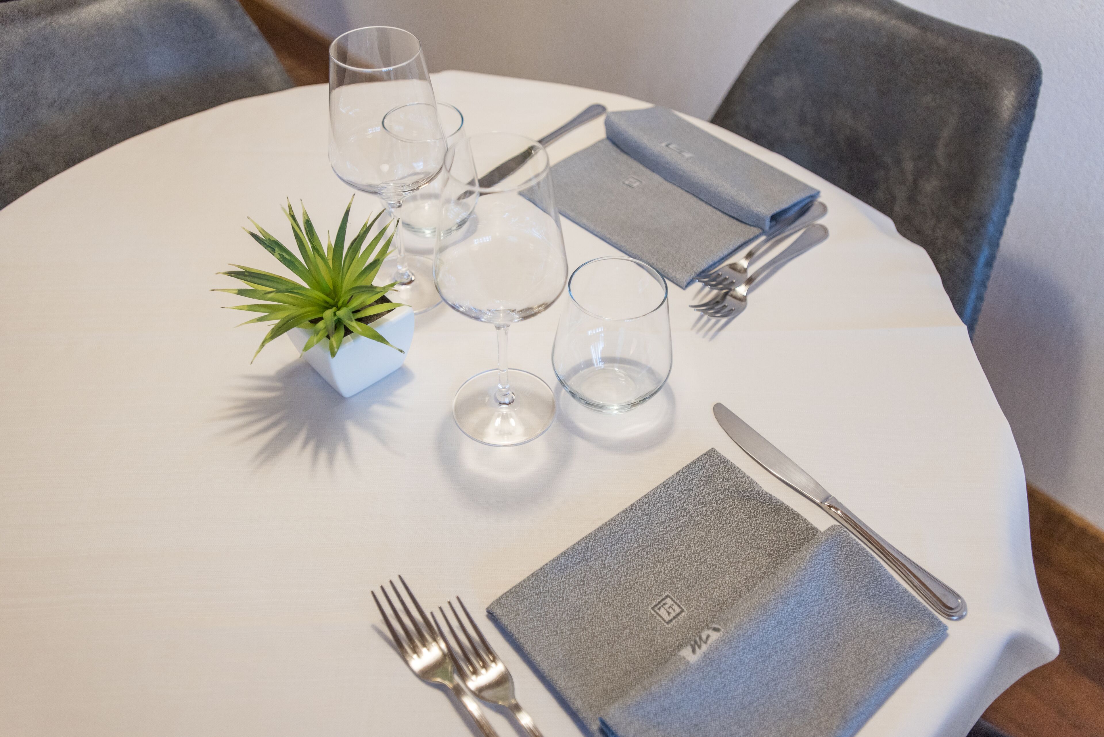 Una mesa redonda de comedor con mantel blanco, una pequeña planta en maceta y cubiertos y vasos ordenados.