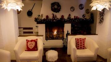Un salón acogedor con dos sillones blancos, un pequeño reposapiés blanco y una mesa de centro de madera. En el centro, una chimenea con un manto decorado con adornos navideños.