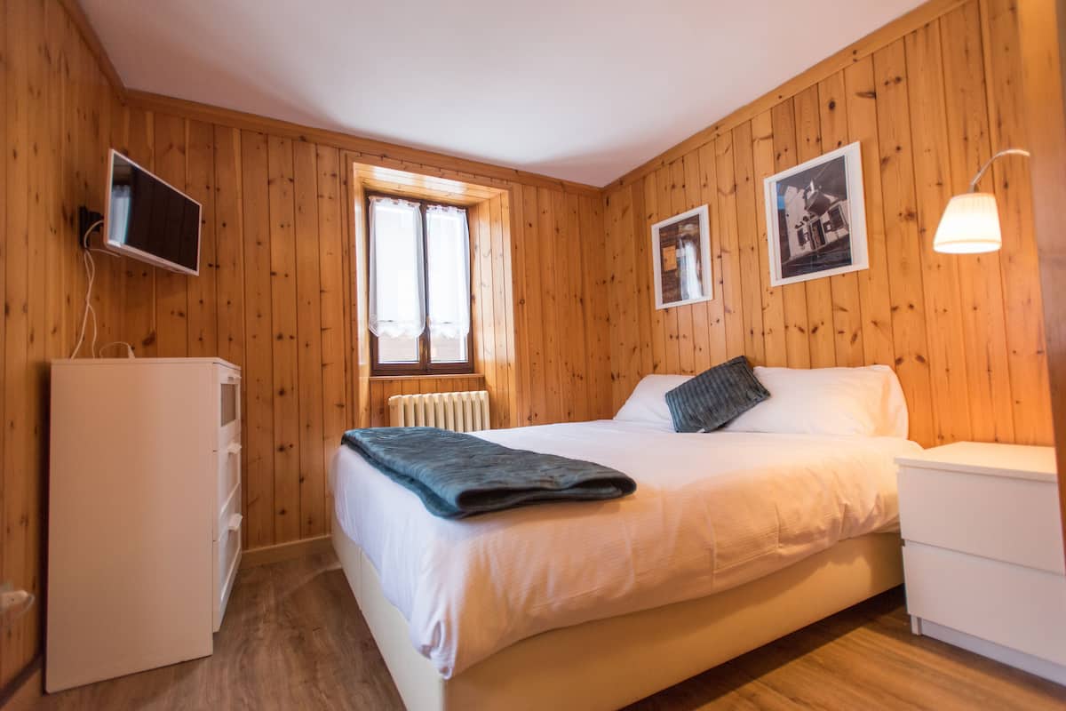A bedroom with wooden walls, a bed with white bedding, a small bedside table, a lamp, a TV on a stand, and a window with curtains.