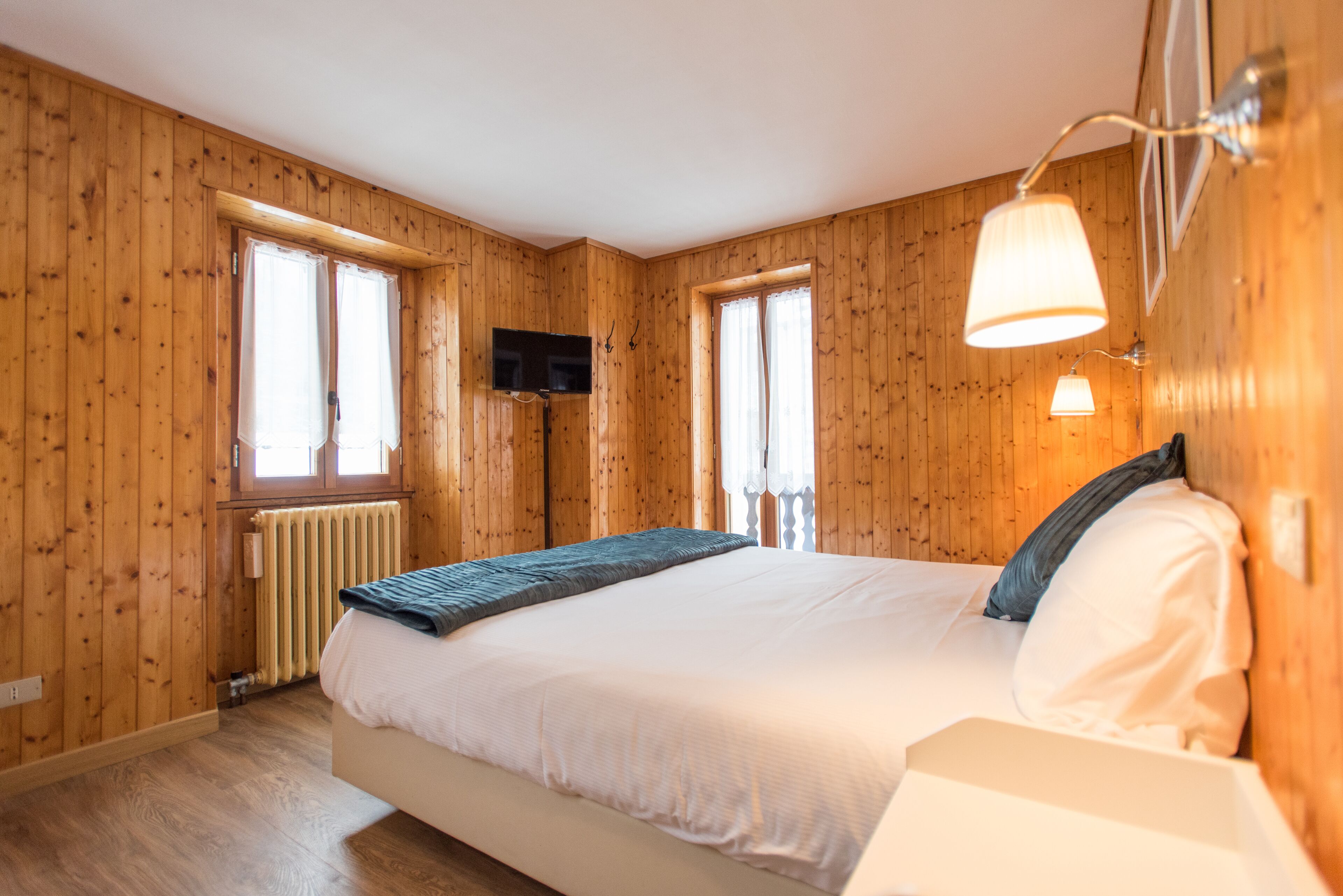 Une pièce aux murs lambrés, comprenant un lit, deux fenêtres, un téléviseur fixé au mur et une lampe de chevet.