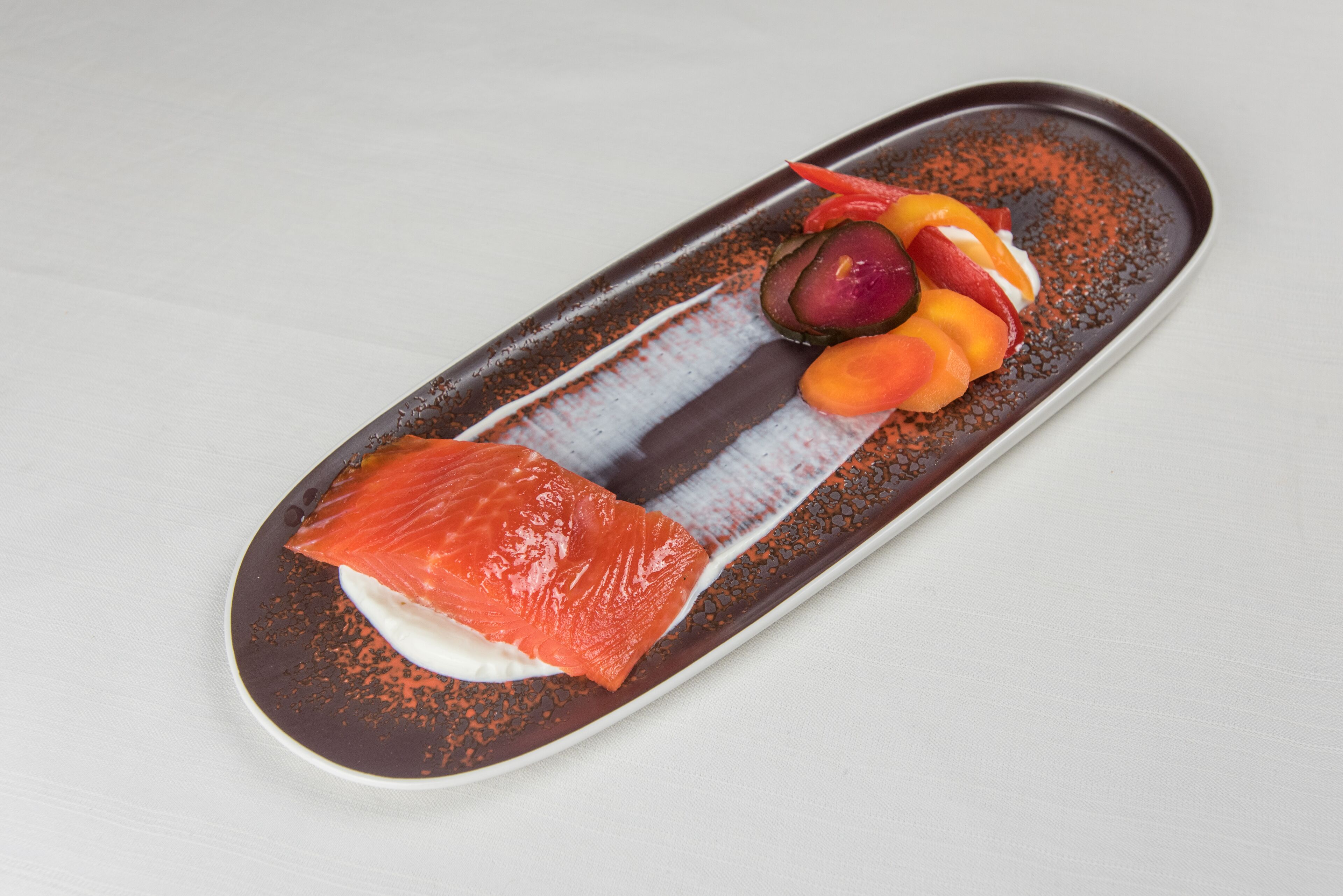 A rectangular plate with a piece of salmon, a dollop of cream, and sliced fruits on a speckled brown plate.