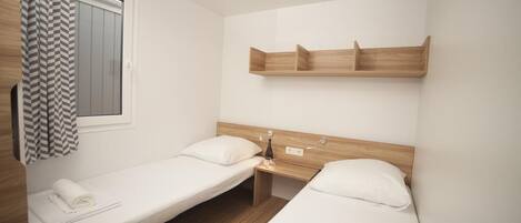 A small, clean room with two single beds, a wooden headboard, a shelf above the beds, and a window with a curtain.