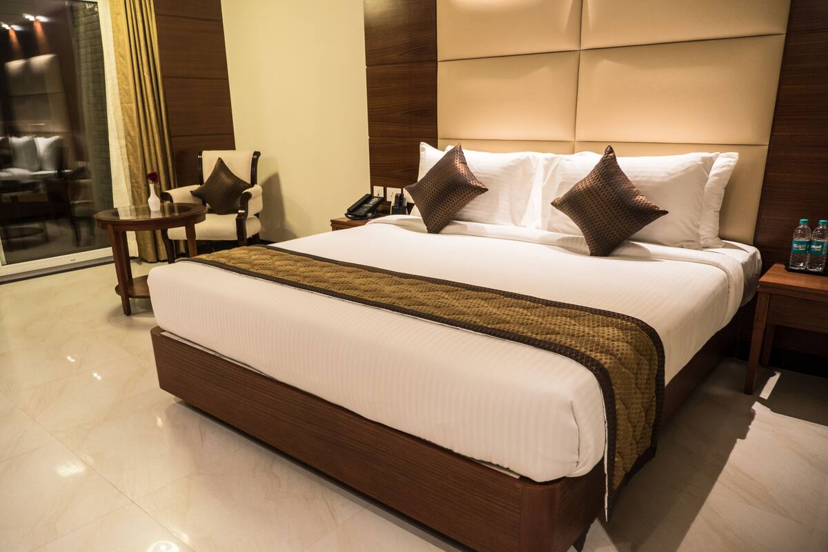 Una cama grande con estructura de madera, ropa de cama blanca y dos almohadones decorativos. Hay una mesita de noche con un teléfono y una botella de agua. Se ve una silla al fondo.
