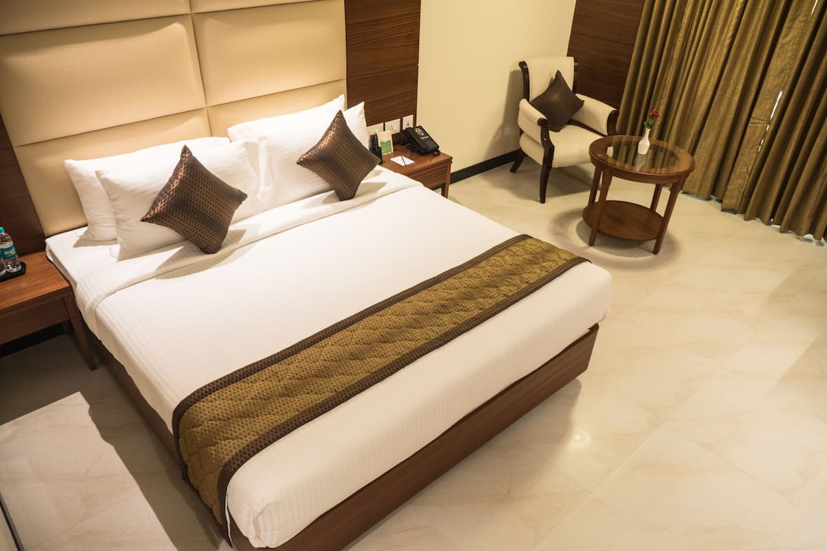 Una cama grande con sábanas blancas y un corredor marrón decorativo, una mesita de noche con un teléfono y una botella, una silla con un jarrón de flores, y una ventana con cortinas.