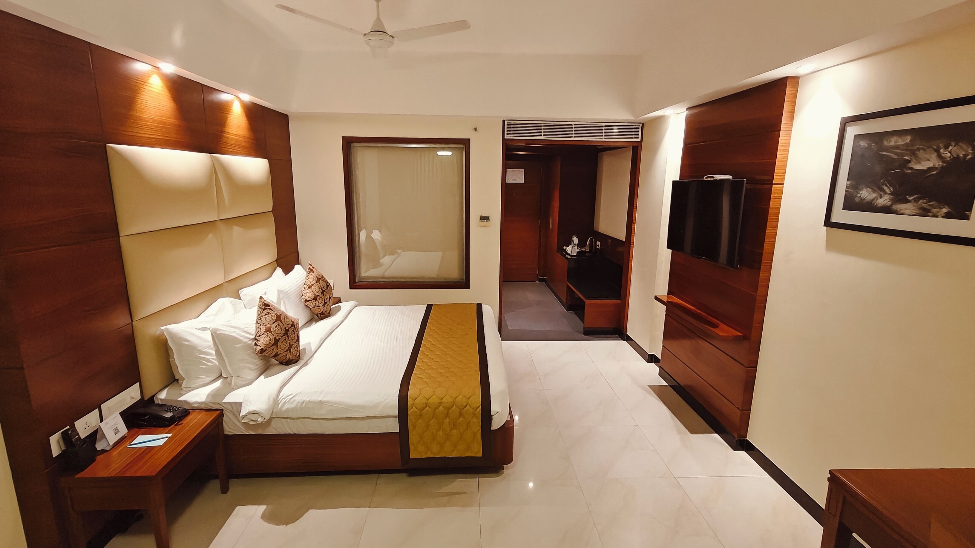 A modern hotel room with a large bed, a bedside table with a phone and papers, a TV mounted on the wall, and a hallway leading to a bathroom.