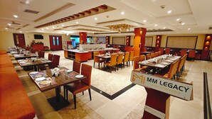 A hotel dining area with multiple tables and chairs, a buffet station, and a sign indicating 'MM Legacy (A unit of MM Hotels)'.