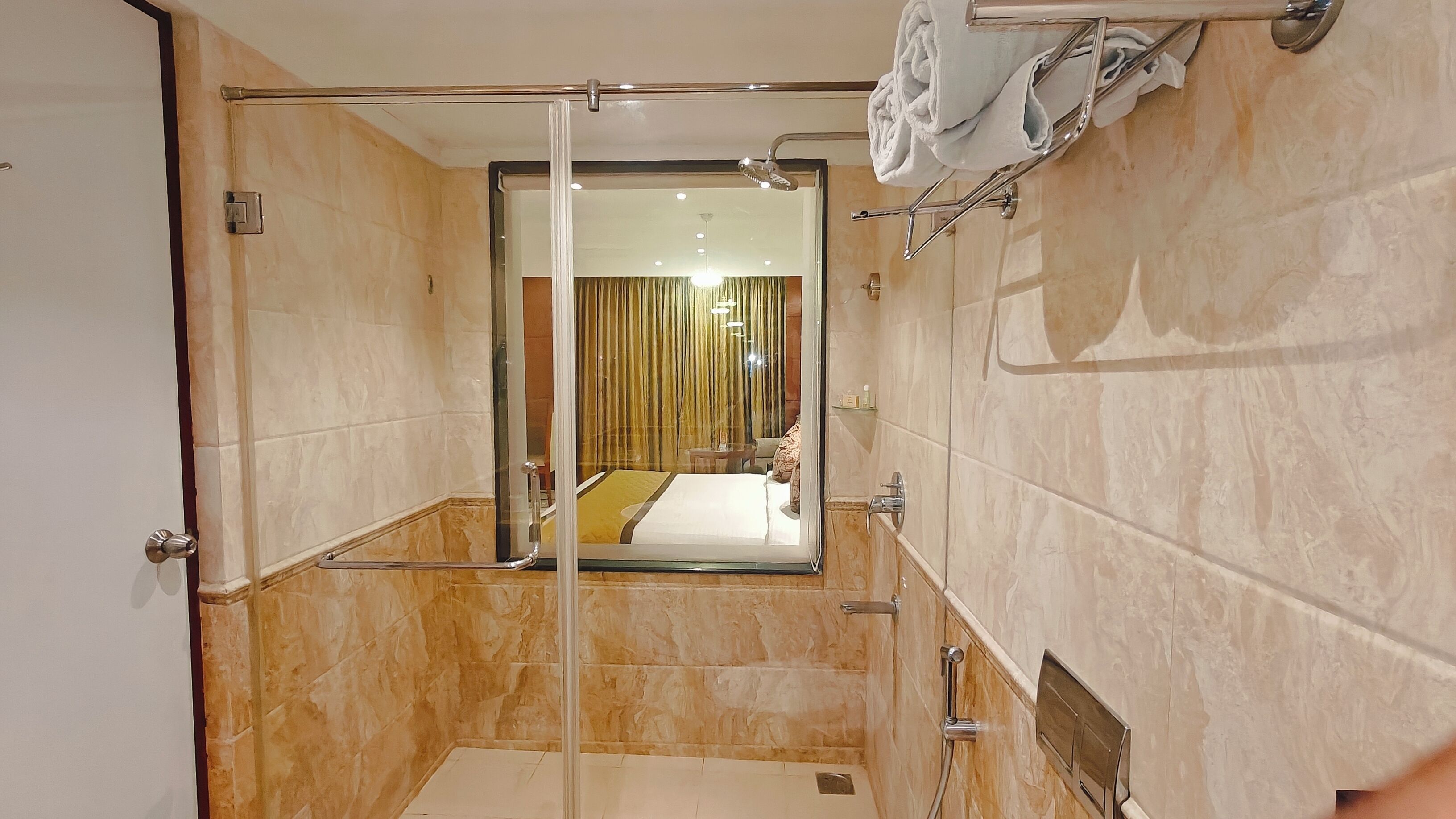 A modern bathroom with a glass shower enclosure, a visible bed in the adjacent room, and a towel rack.