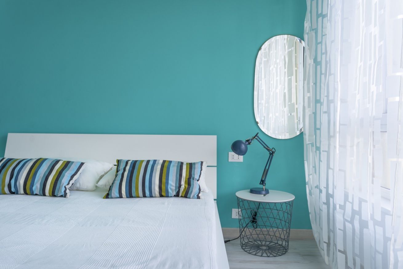 Une chambre à coucher avec un lit, des coussins à rayures, une petite table ronde avec une lampe, et une fenêtre avec des rideaux à motifs.