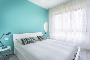A bedroom with a teal wall, a white bed, striped pillows, a bedside table with a lamp, and a window with sheer curtains.