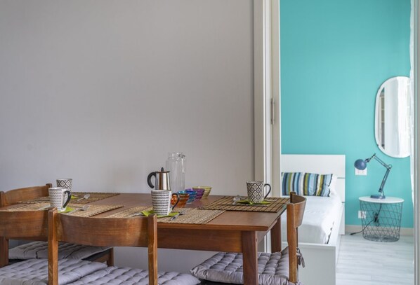 Een eetruimte met een houten tafel voor vier personen, omringd door stoelen met kussens. Een bed met een gestreept kussen is op de achtergrond zichtbaar.