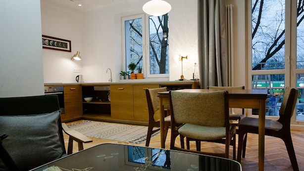 A modern kitchen with wooden cabinets, a dining table, and chairs. There is a window with a view of buildings and trees. - Warsaw Concierge Saska Studio (Warsaw)