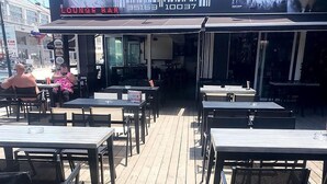 An outdoor seating area with tables and chairs, a digital signboard, and a view of the street.