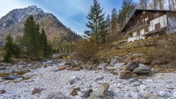 Eine Bergingerhütte mit Steinfundament, umgeben von einem felsigen Bach und bewaldetem Gebiet.