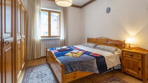 Ein Schlafzimmer mit einem hölzernen Bett, einem Nachttisch, einer Lampe, einem Fenster mit Vorhängen und einer Heizung.