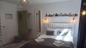 Un dormitorio con cama blanca, almohadas grises y blancas, una manta gris y una toalla doblada sobre la cama. Se ve una puerta de madera con un teléfono, un espejo en la pared y un vinilo con el horizonte de una ciudad sobre la cama.