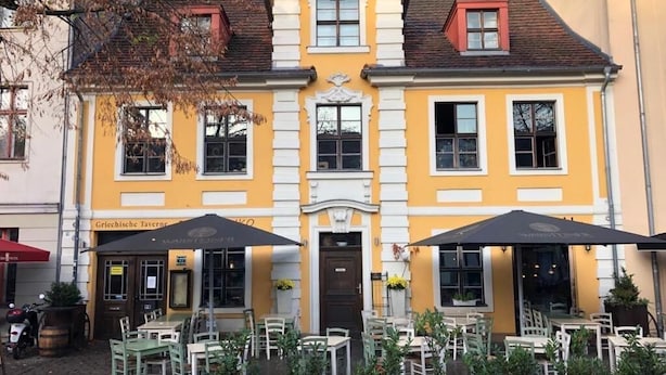 Un edificio amarillo con una puerta central, dos zonas de estar al aire libre con sombrillas y plantas en macetas.