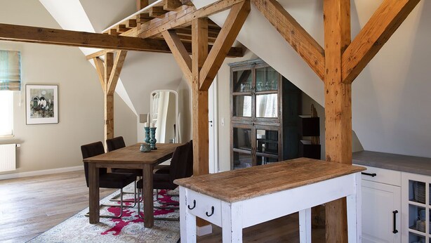 Un comedor con una mesa y sillas de madera, un armario blanco y una escalera con vigas de madera a la vista.