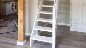 Una escalera de madera blanca en una habitación con vigas de madera, armarios blancos y un techo visible con tragaluces.