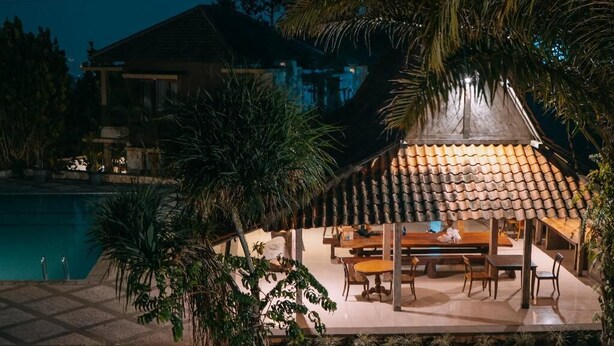 Un patio con piscina, rodeado de palmeras y una zona de estar cubierta con mesas y sillas.
