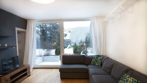 A modern living room with a sectional sofa, a flat-screen TV on a wooden stand, and a large sliding glass door leading to a balcony. - Warsaw Concierge Family Apartment (Warsaw)