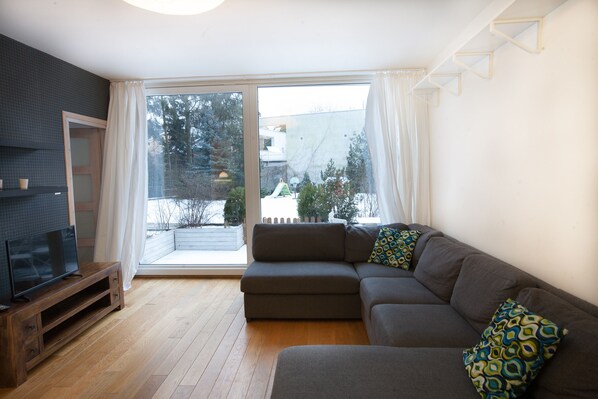 A modern living room with a sectional sofa, a flat-screen TV on a wooden stand, and a large sliding glass door leading to a balcony.