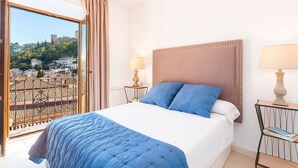 A hotel room with a large bed, two bedside lamps, a view of a cityscape through a balcony, and a window with curtains.