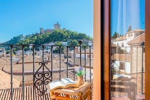 Un balcón con vista a un castillo histórico en la cima de una colina, una silla de mimbre y un libro abierto.