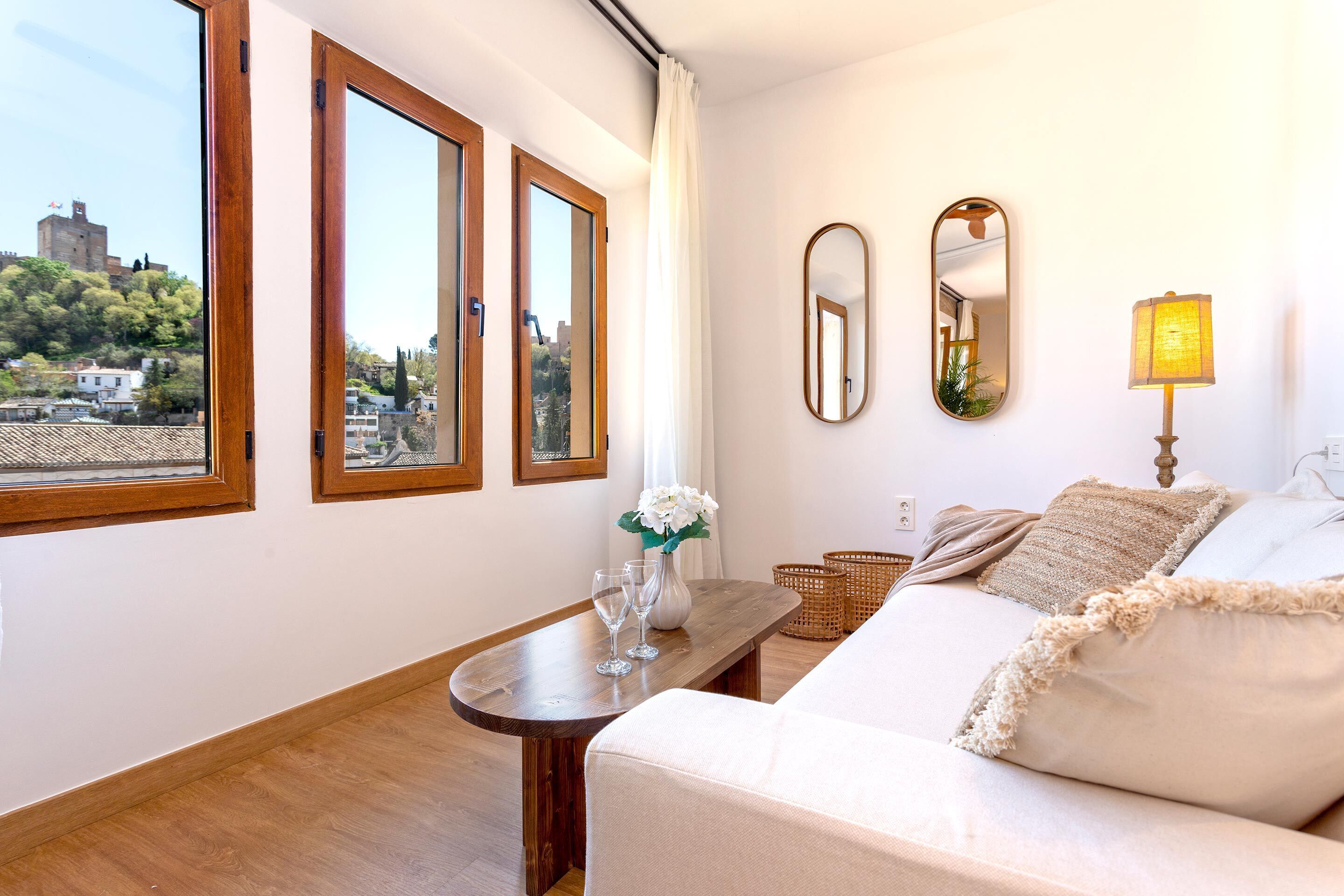 A room with a wooden floor, a sofa, a table with a vase and glasses, and two mirrors on the wall.