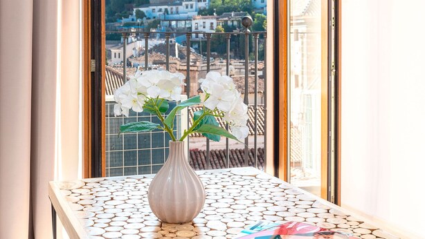 A table with a vase and a magazine, a window with a view of buildings and trees, and a curtain on the left.