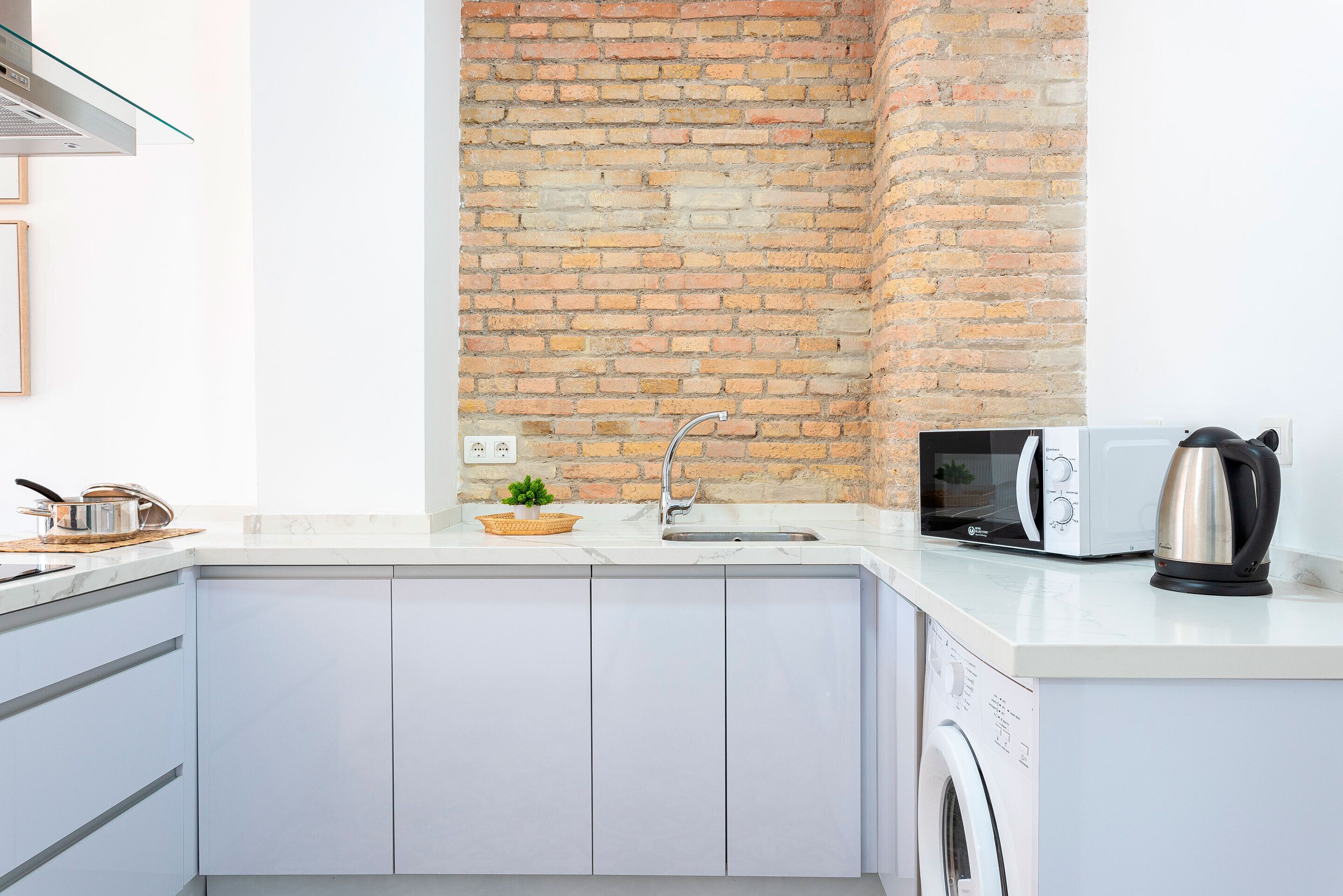 Een moderne keuken met een bakstenen muur, witte kasten, een ingebouwde magnetron en een waterkoker op het aanrecht.