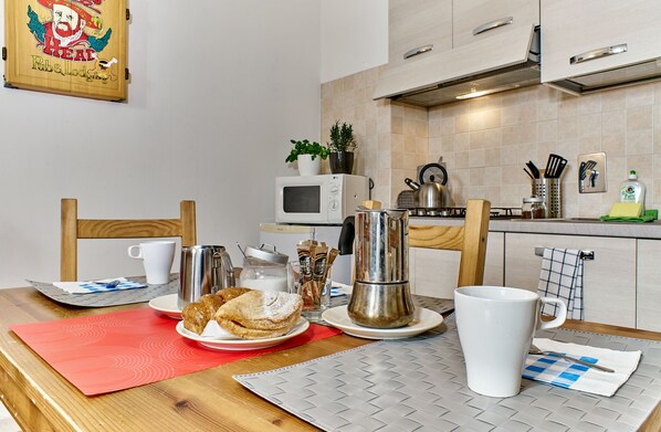 Una cocina equipada con microondas, tostadora, hervidor y varios utensilios. Hay una mesa con platos, tazas y un mantel individual rojo.