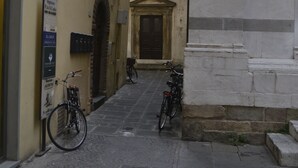 A narrow cobblestone street with bicycles parked on the side, a building with a signboard, and a door leading to a building with a stone facade. - Bilocale Torre delle Ore (Lucca)