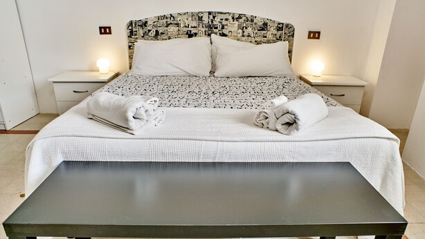A neatly made bed with a patterned bedspread and pillows, two bedside tables with lamps, and a bench in front of the bed. - Bilocale Torre delle Ore (Lucca)