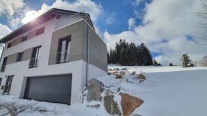A modern two-story house with a garage, surrounded by snow-covered ground and trees.