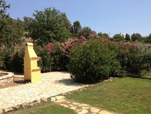 A yellow outdoor structure with a stone pathway leading to it, surrounded by green bushes and pink flowers.