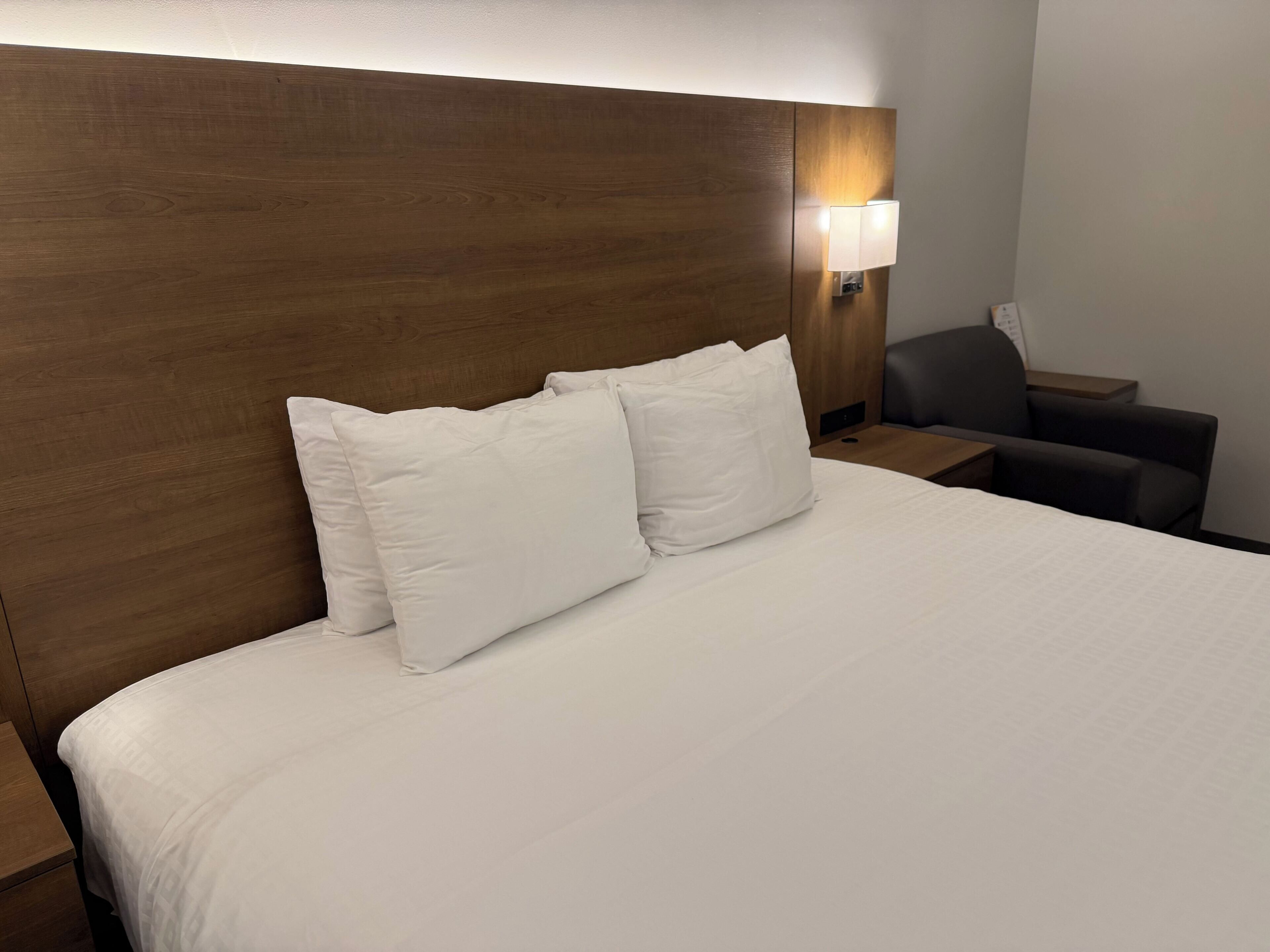 Een hotelkamer met een houten hoofdeinde, een bed met witte beddengoed, een nachtkastje, een lamp en een stoel.