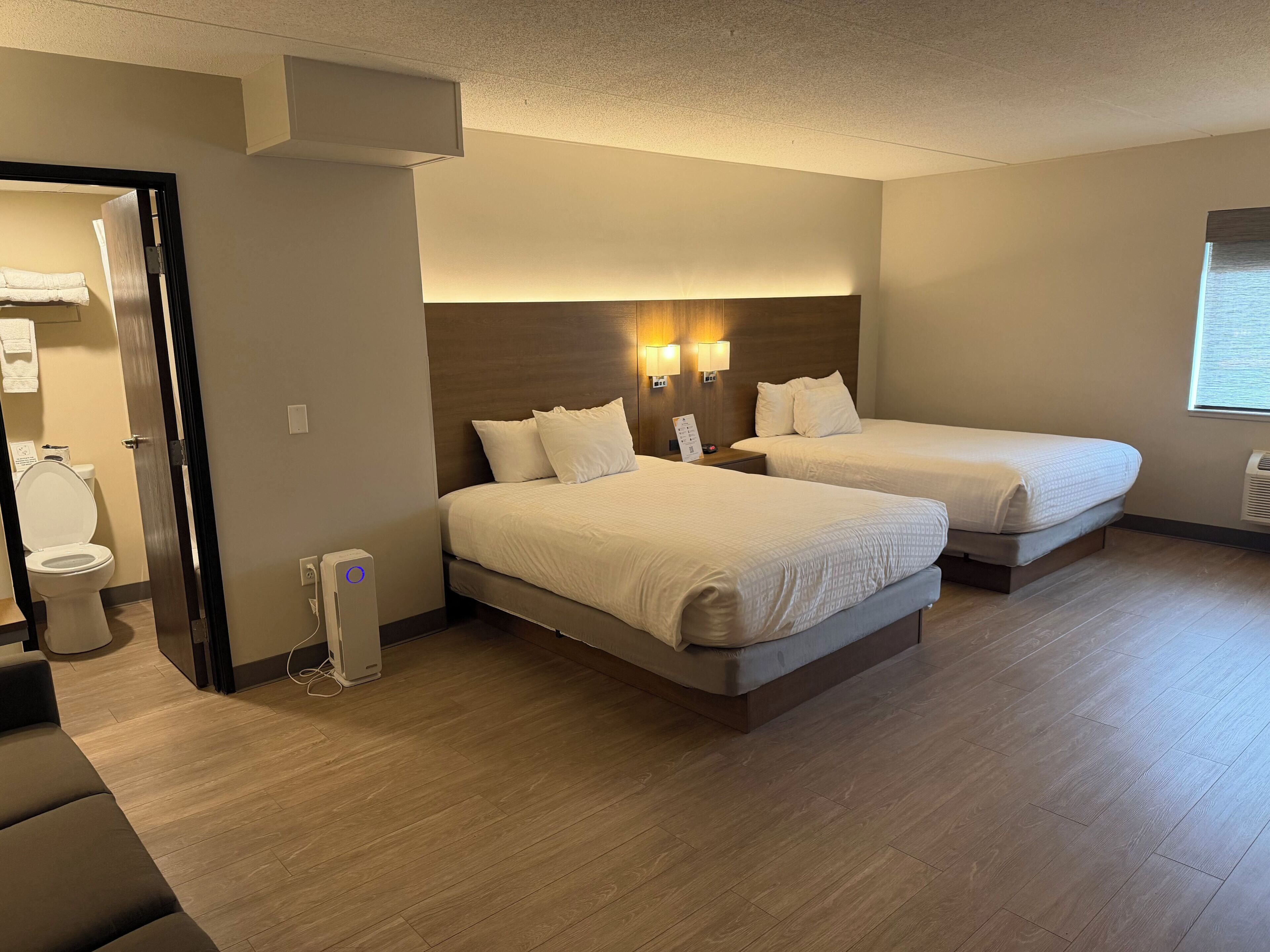 A hotel room with two beds, a wooden floor, a wall-mounted heater, and a window with blinds.