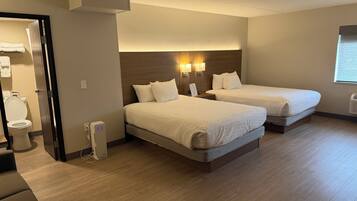 A hotel room with two beds, a wooden floor, a wall-mounted heater, and a window with blinds.