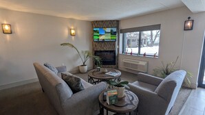 Una sala de estar con chimenea, un televisor de pantalla plana montado encima y un gran ventanal con vistas al exterior.