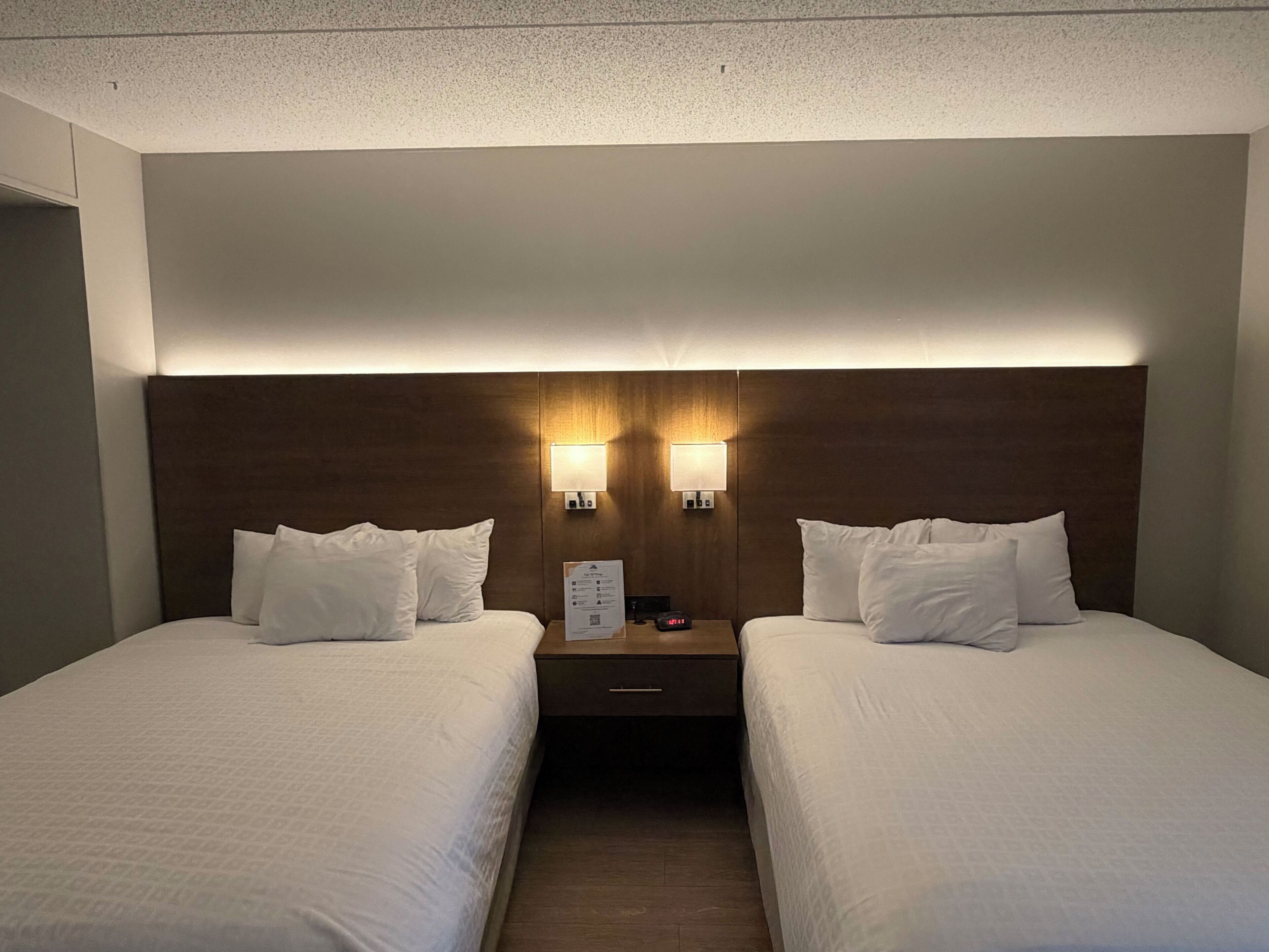 Habitación de hotel con dos camas, cabecero de madera, dos lámparas de pared y una mesita de noche con un reloj.