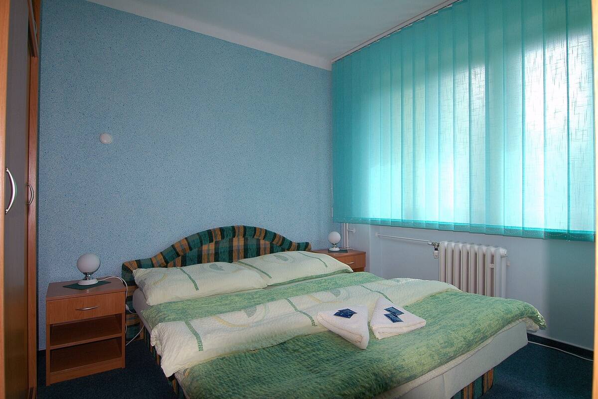 A room with a bed, two bedside tables, a window with blinds, and a radiator.
