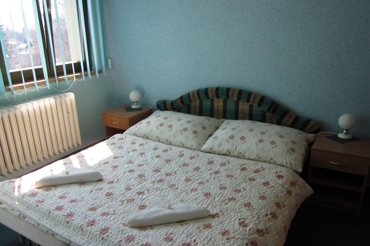 A bedroom with a bed, two bedside tables, a radiator, and a window with blinds.