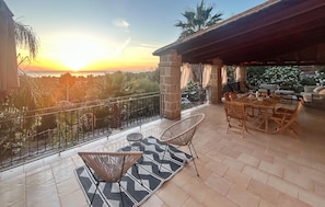 A terrace with a table and chairs, a view of the sunset, and a covered area with seating.