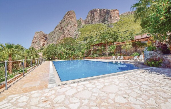 A swimming pool with stone flooring, surrounded by palm trees and a rocky mountain backdrop.