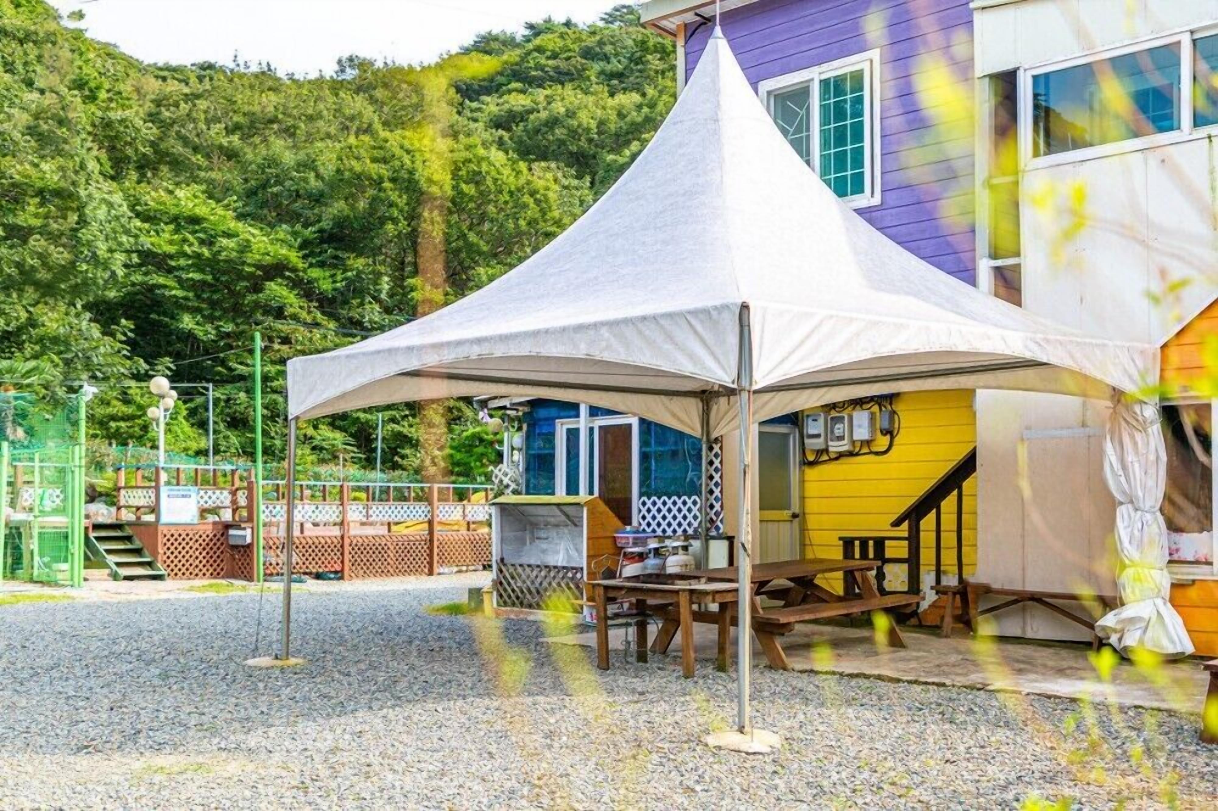 Un espace extérieur aménagé avec une tente, une table de pique-nique et des bancs, un escalier menant à un bâtiment à la façade violette et jaune, et une colline boisée en arrière-plan.