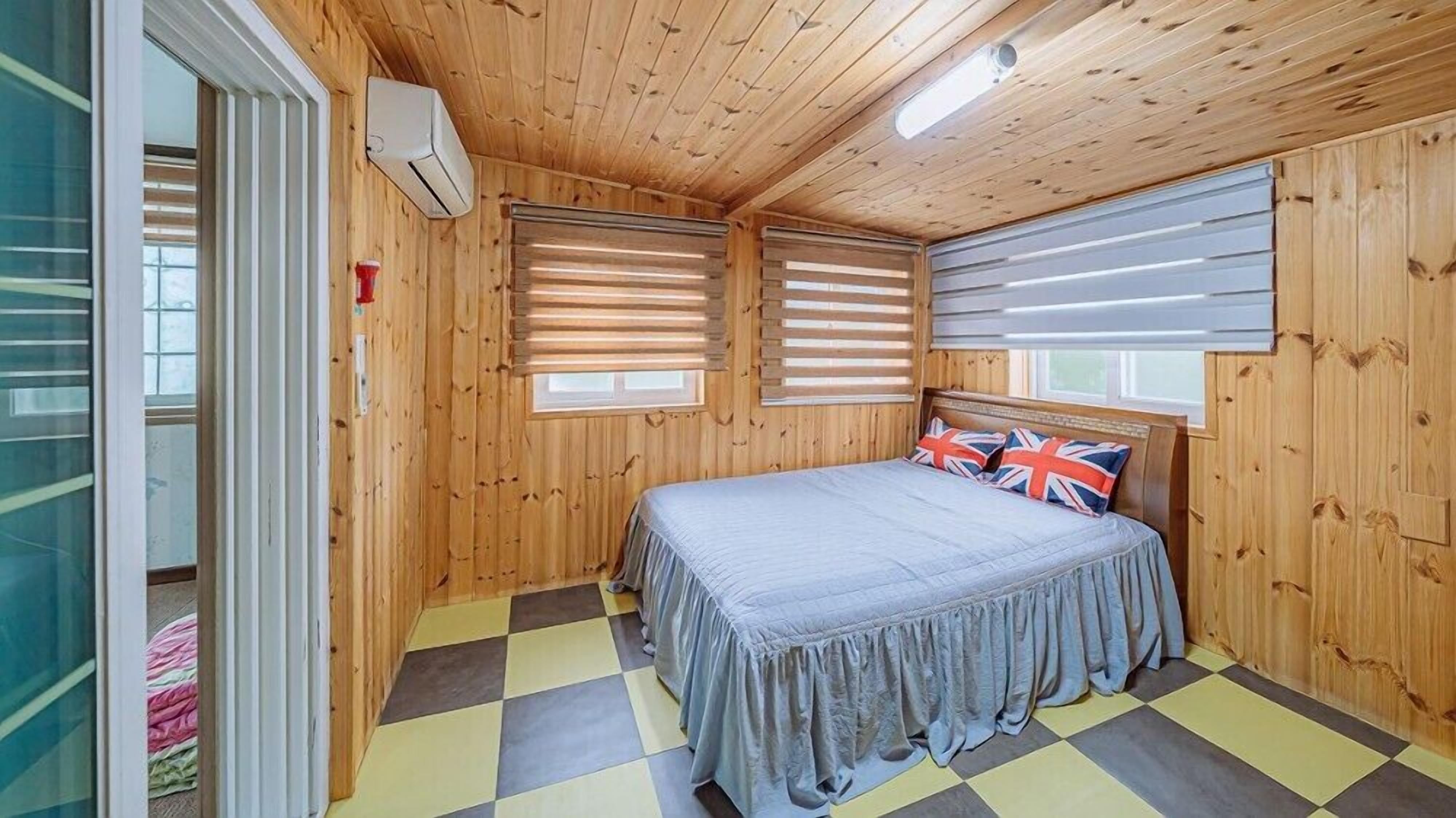 A wooden-paneled room with a bed, two windows with blinds, a ceiling light, and a checkered floor.