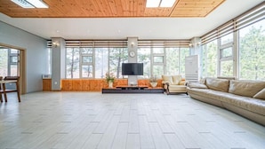 A spacious living room with a large TV, comfortable seating, and large windows offering a view of trees. - Sunny Hill Pension (Gapyeong)