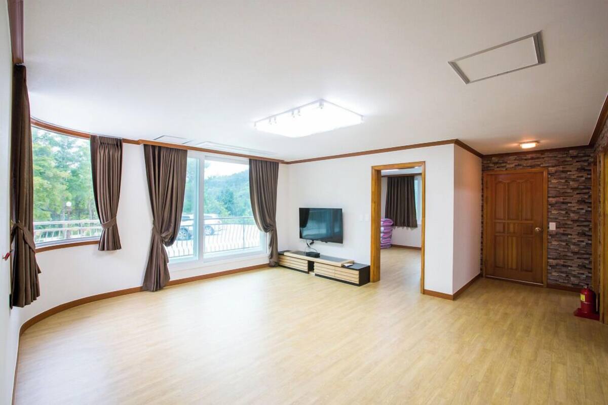 A spacious living room with a large window, a flat-screen TV, and a wooden floor.