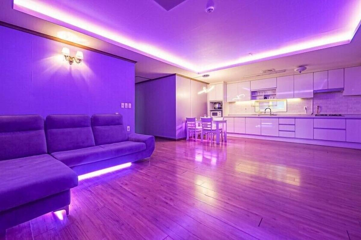 A modern living room with a purple-lit sofa, a dining area with chairs, and a kitchen with white cabinets.